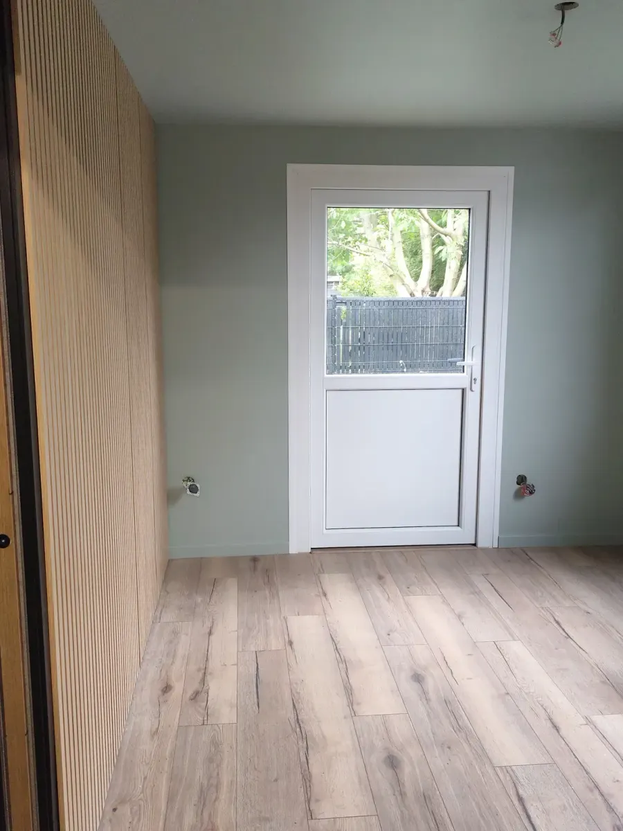 Bureau après travaux : murs gris-vert clair, sol stratifié chêne, mur habillé de tasseaux de bois, porte vitrée blanche au fond