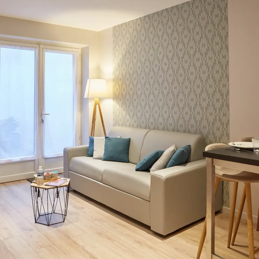 Salon dont un mur est tendu d’un papier peint à motif damassé gris, derrière un canapé clair