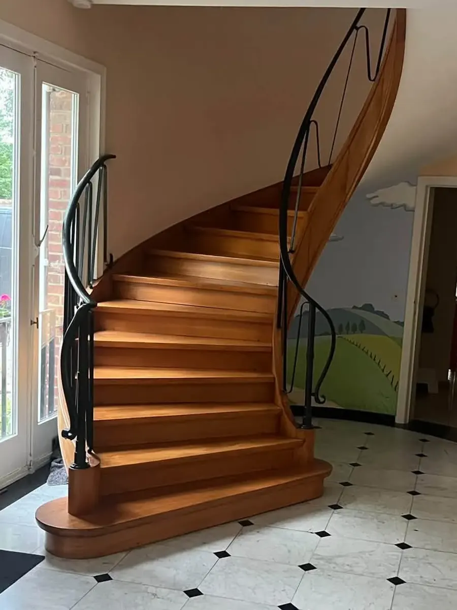 Escalier tournant en bois verni avant travaux, rampe en fer forgé noire et murs rose pâle