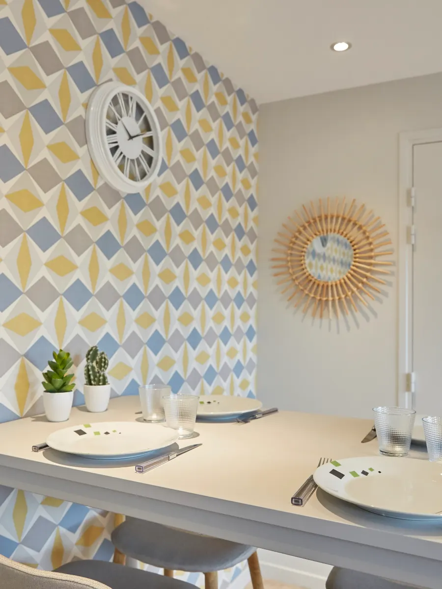 Coin repas dont le mur et le rampant sont habillés d’un papier peint géométrique jaune, bleu et gris