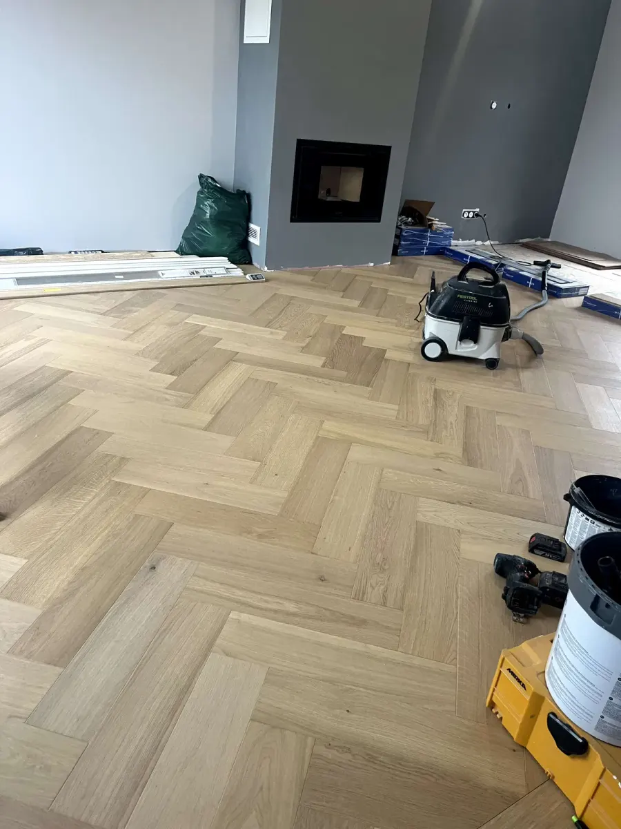 Parquet en chevron chêne clair fraîchement posé, mur gris et foyer encastré au fond