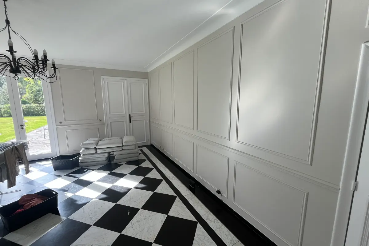 Grande pièce de réception aux murs à panneaux blancs, lustre en fer forgé et baie donnant sur un jardin