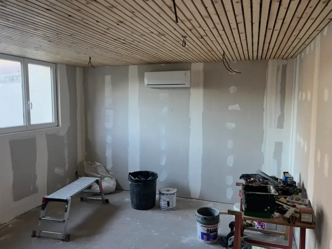 Pièce en travaux : cloisons de plâtre brut avec bandes apposées, plafond en lames de bois clair, escabeau et seaux au sol